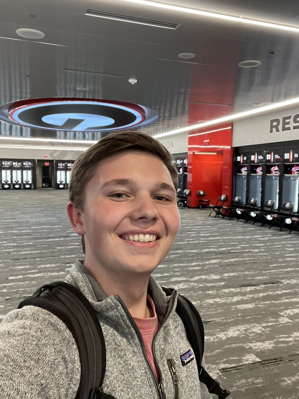 Ethan Wallace smiling, wearing a gray fleece and backpack, inside a University of Georgia athletics facility